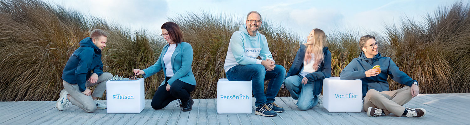 Fünf Personen vom NORDkind Team sitzen oder hocken lächelnd auf Holzboden verteilt um drei weiße Würfel mit Beschriftung "Plietsch", "Persönlich", "Von Hier". Auf dem linken Würfel spielen zwei Personen Schach. Dominik, der Inhaber, sitzt auf dem mittleren Würfel. Zwei Personen lehnen am rechten Würfel, einer von ihnen isst ein Fischbrötchen. Im Hintergrund blauer Himmel und hohes Gras.