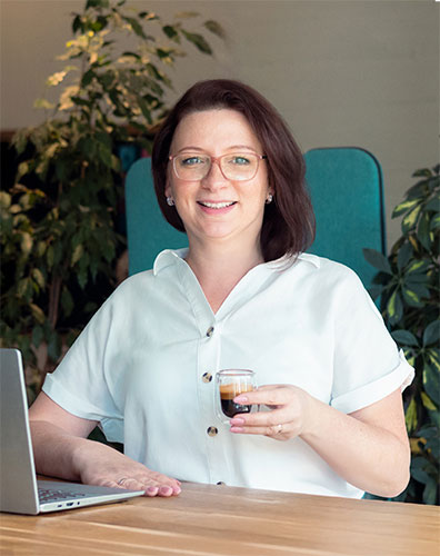 Nina sitzt am Schreibtisch vor dem Laptop mit einem Glas Espresso in der Hand und lächelt. Sie trägt eine weiße Bluse und eine Brille.
