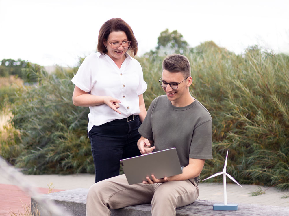 Julian sitzt mit Laptop auf dem Schoß auf einem Holzstamm, neben ihm steht ein Modell von einem Windrad. Er lächelt und zeigt auf den Laptop. Hinter ihm steht Nina und zeigt auch lächelnd auf den Laptop.