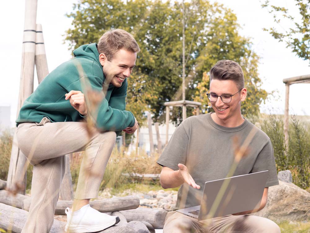 Julian und Paul sitzen draußen auf Baumstämmen und schauen lächelnd auf den Laptop.