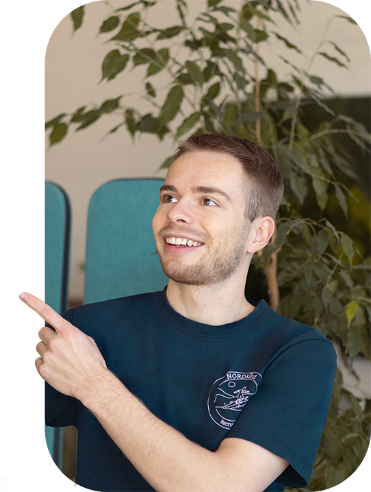 Paul lächelt und zeigt mit dem Finger links aus dem Bild heraus. . Er trägt ein dunkelblaues T-Shirt.